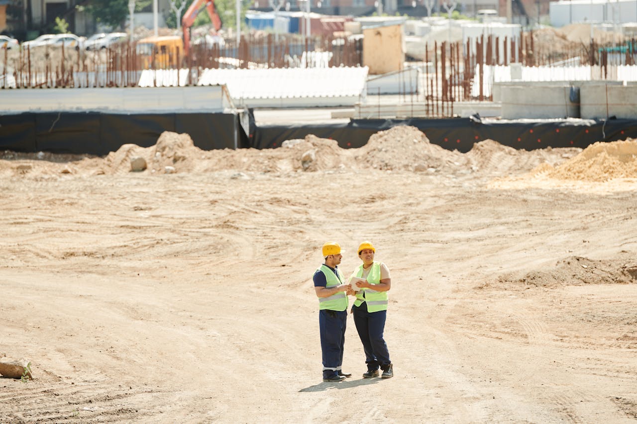 Two workers discussing plans on a sandy construction site, wearing safety gear.